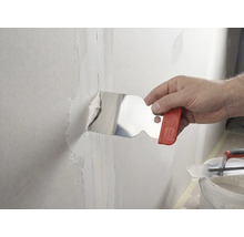 En hand applicerar spackel på en väggfog med en spackelspade i rostfritt stål.