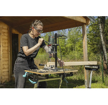 En kvinna borrar i en träbalk med ett borrstativ och en borrmaskin på en mobil arbetsbänk i en trädgård.
