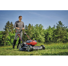 En man klipper en grön gräsmatta med en Einhell batteridriven gräsklippare i en solig trädgård med träd i bakgrunden.
