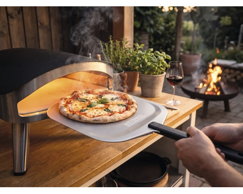 En nybakad pizza tas ut ur en bärbar pizzaugn på ett träbord i trädgården med en pizzaspade med Tenneker Logo.