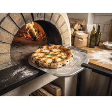 Nybakad pizza på en pizzaspade med Tenneker Logo framför en brinnande stenugn i ett rustikt kök.