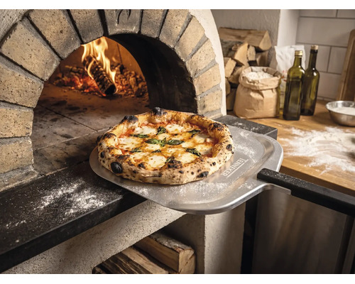 Nybakad pizza på en pizzaspade med Tenneker Logo framför en brinnande stenugn i ett rustikt kök.