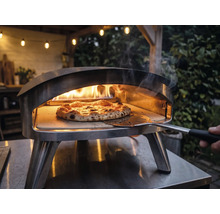 Pizzaugn i rostfritt stål för utomhusbruk med brinnande lågor och en pizza på en pizzasten som flyttas med en pizzaspade.