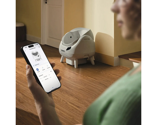 En person styr en självrengörande Catlink kattlåda via en app på en smartphone i ett ljust rum.