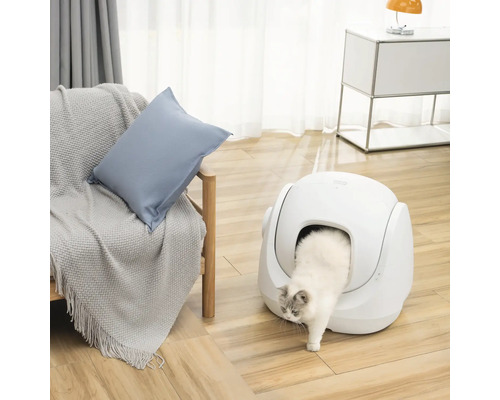 Vit katt som kliver ur en modern, självrengörande kattlåda i ett ljust vardagsrum med trägolv och fåtölj.