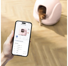 Smartphone-applikation för styrning av en självrengörande kattlåda, i bakgrunden syns enheten med en katt på ett trägolv.