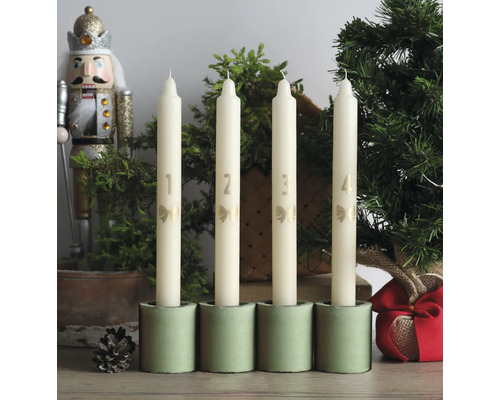 Fyra cremefärgade adventsljus med siffrorna ett till fyra och rosetter i gröna hållare, bredvid en nötknäppare och små julgranar.