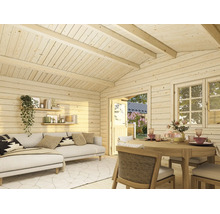 Interiörvy av en ljus trädgårdsstuga i trä med vardagsrum, soffa, matbord och utsikt över trädgården genom dörr och fönster.