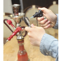 En hand tänder vattenpipskol med en gasbrännare, i bakgrunden står en vattenpipa på ett bord.