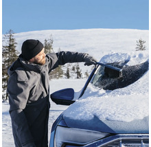 Man tar bort snö från vindrutan på en bil med en snöborste i ett snöigt vinterlandskap.
