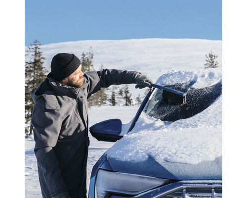 Man tar bort snö från vindrutan på en bil med en snöborste i ett snöigt vinterlandskap.