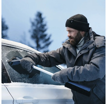 En man i vinterkläder tar bort snö från en bilruta med en isskrapa.