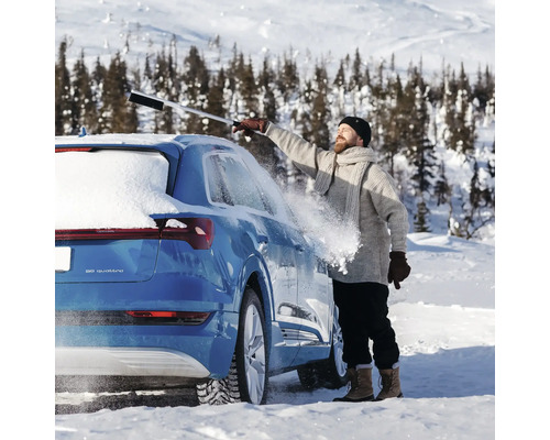 En man tar bort snö från en bil med en isskrapa med borste i ett snöigt landskap.