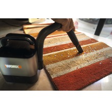 En sladdlös dammsugare används på en randig matta för att ta bort små partiklar. Worx företagslogotyp.