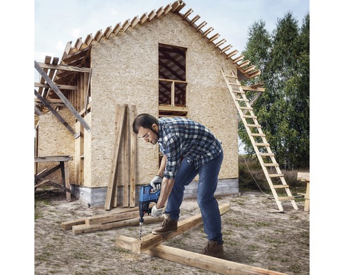Hantverkare borrar ett hål med en borrmaskin i en träbjälke framför ett hus under konstruktion.