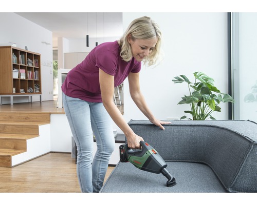 Kvinna dammsuger en grå soffa med en batteridriven dammsugare i ett ljust vardagsrum.