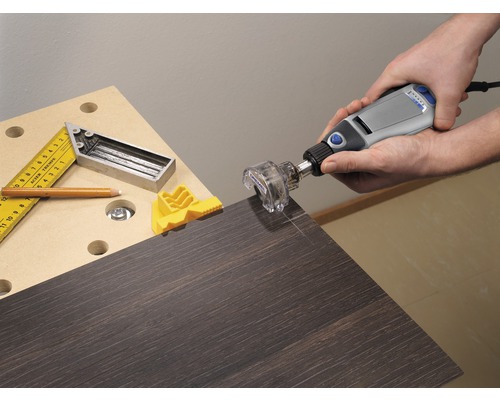 En person använder ett Dremel multifunktionsverktyg med ett frästillsats på en mörk träskiva. En vinkelhake, en penna och en gul arbetsstyckesspännare ligger på arbetsbänken.