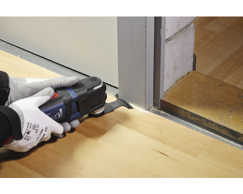 En person använder ett multifunktionsverktyg för att såga ett trägolv vid en dörr.