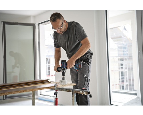 En man borrar ett hål i en träplanka med en sladdlös borrmaskin. Han bär skyddsglasögon och arbetskläder.