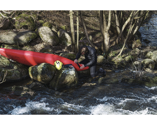 Man reparerar kajak vid flodstrand med stenar och träd.