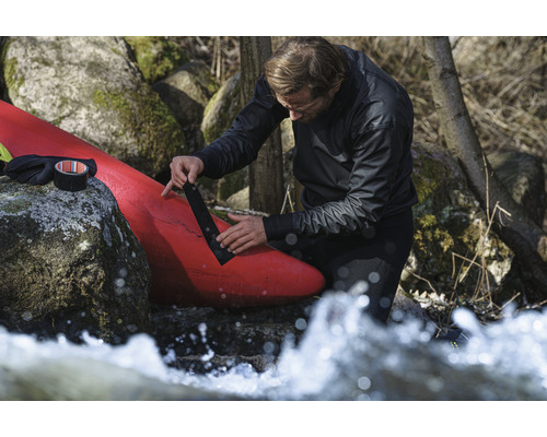 En man reparerar en röd kajak med svart tejp nära stenar och rinnande vatten.