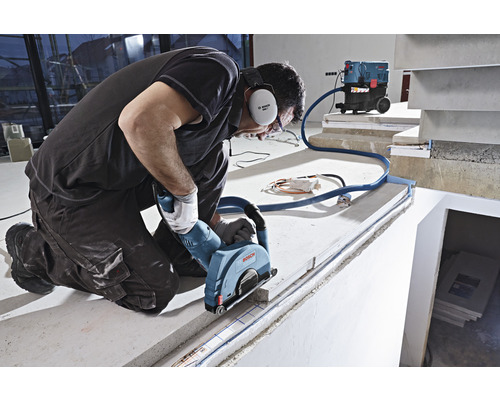 En man knäböjer och skär en fog i en betongyta med en väggspårfräs, medan en dammsugare suger upp dammet.