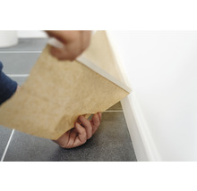 Person installerar en golvlist med pappersunderlag längs en vägg på ett klinkergolv.