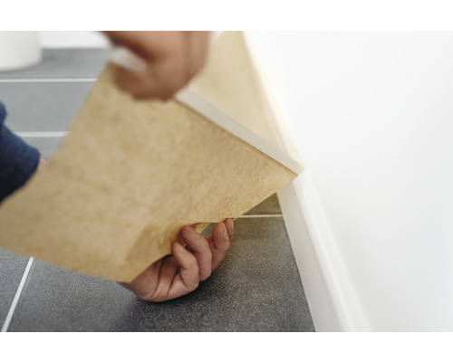 Person installerar en golvlist med pappersunderlag längs en vägg på ett klinkergolv.