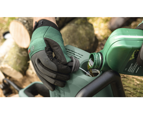 En person fyller på olja i en motorsåg. I bakgrunden syns vedträn.