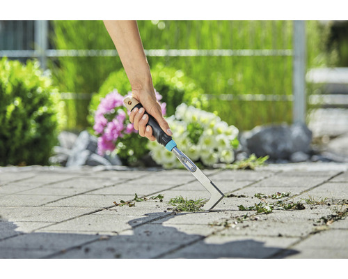 En person tar bort ogräs från gatstenar i en trädgård med en fogskrapa.