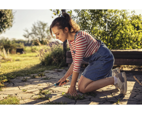 Kvinna tar bort ogräs från stenläggning i trädgården med ett ogräsjärn.