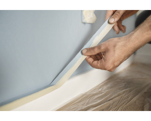 En person fäster maskeringstejp på en vägg som förberedelse för målningsarbeten.