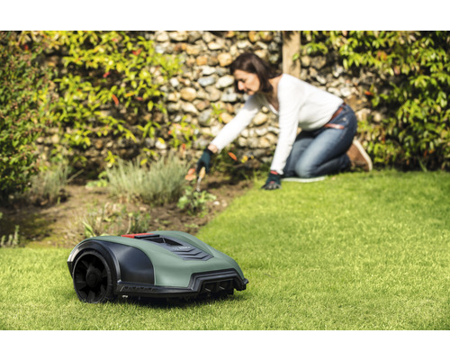 En robotgräsklippare står på en gräsmatta, medan en kvinna arbetar i trädgården i bakgrunden.