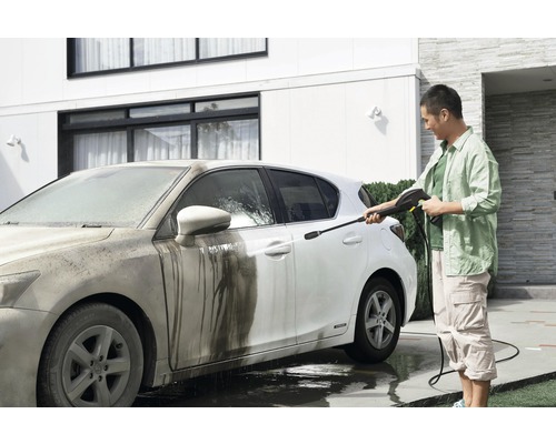 Man rengör en smutsig bil med en högtryckstvätt framför ett hus.