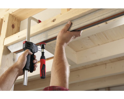 En hand fäster en metallskena med en skruvtving på en träkonstruktion.