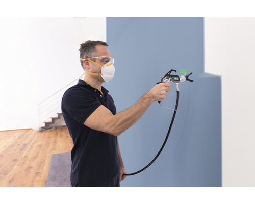 Man målar väggen med en färgspruta, han bär skyddsmask och skyddsglasögon