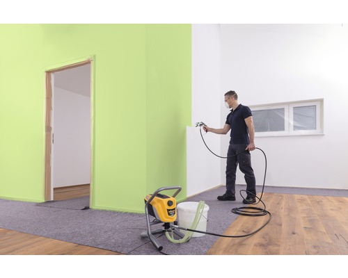 En man sprutar färg med en färgspruta i ett rum. I förgrunden står en färgspruta.