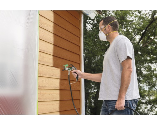 Man målar husvägg med färgspruta och bär skyddsmask och skyddsglasögon.