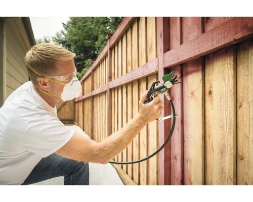 Man målar trästaket med färgspruta, med skyddsglasögon och andningsmask.