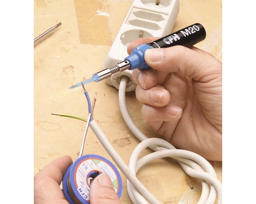 En hand löder en elektrisk ledning med en lödkolv. En spole med lödtenn och ett grenuttag ligger på bordet.