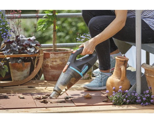 En person använder en sladdlös handdammsugare på en balkong för att ta bort löv. Blomkrukor och växter syns i bakgrunden.