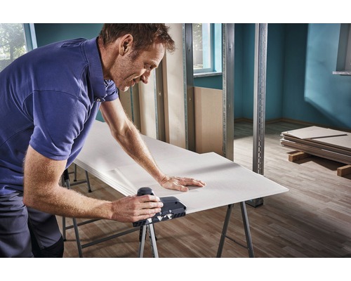 Man bearbetar gipsskivor med en kantbårshyvel på en arbetsbänk i ett råbygge.