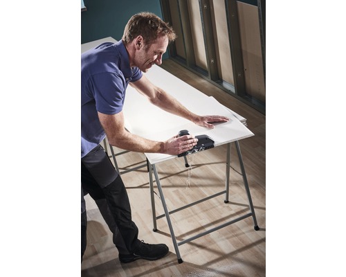 En man skär en gipsskiva med ett verktyg på ett arbetsbord.