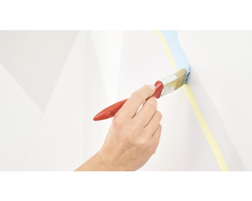 En hand målar med en pensel blå färg längs en gul maskeringstejp på en vägg.