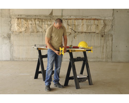 En man arbetar med en Stanley sågbock i en byggmiljö. På sågbocken ligger ett vattenpass, ett måttband och en bygghjälm.