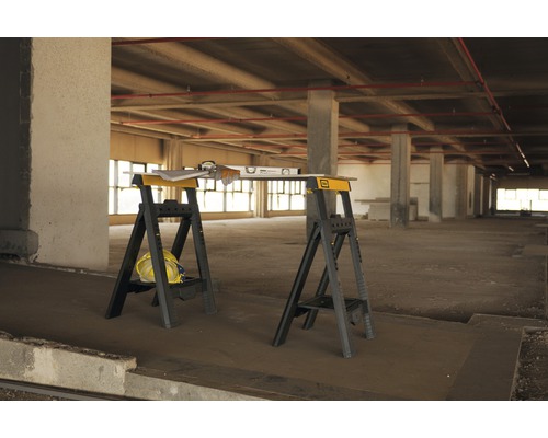 Två svarta och gula sågbockar med vattenpass, handskar, byggnadsplan och bygghjälm i ett råbygge.