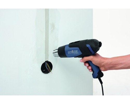 Varmluftspistol i handen framför en vägg med exponerade kablar