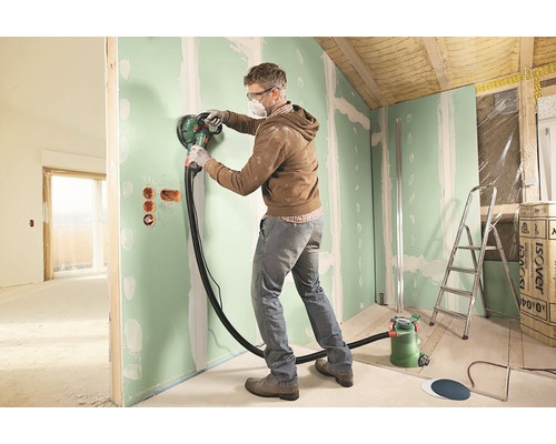 En man slipar en vägg i ett rum med en takslipmaskin.