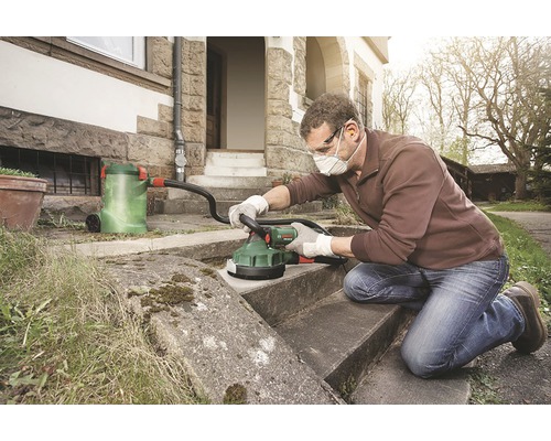 En man rengör stentrappor med en betongslipmaskin framför ett hus. Slipmaskinen är ansluten till en dammsugare.