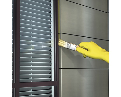 Utomhusbild av en person med handskar som målar en vägg bredvid ett fönster med persienner